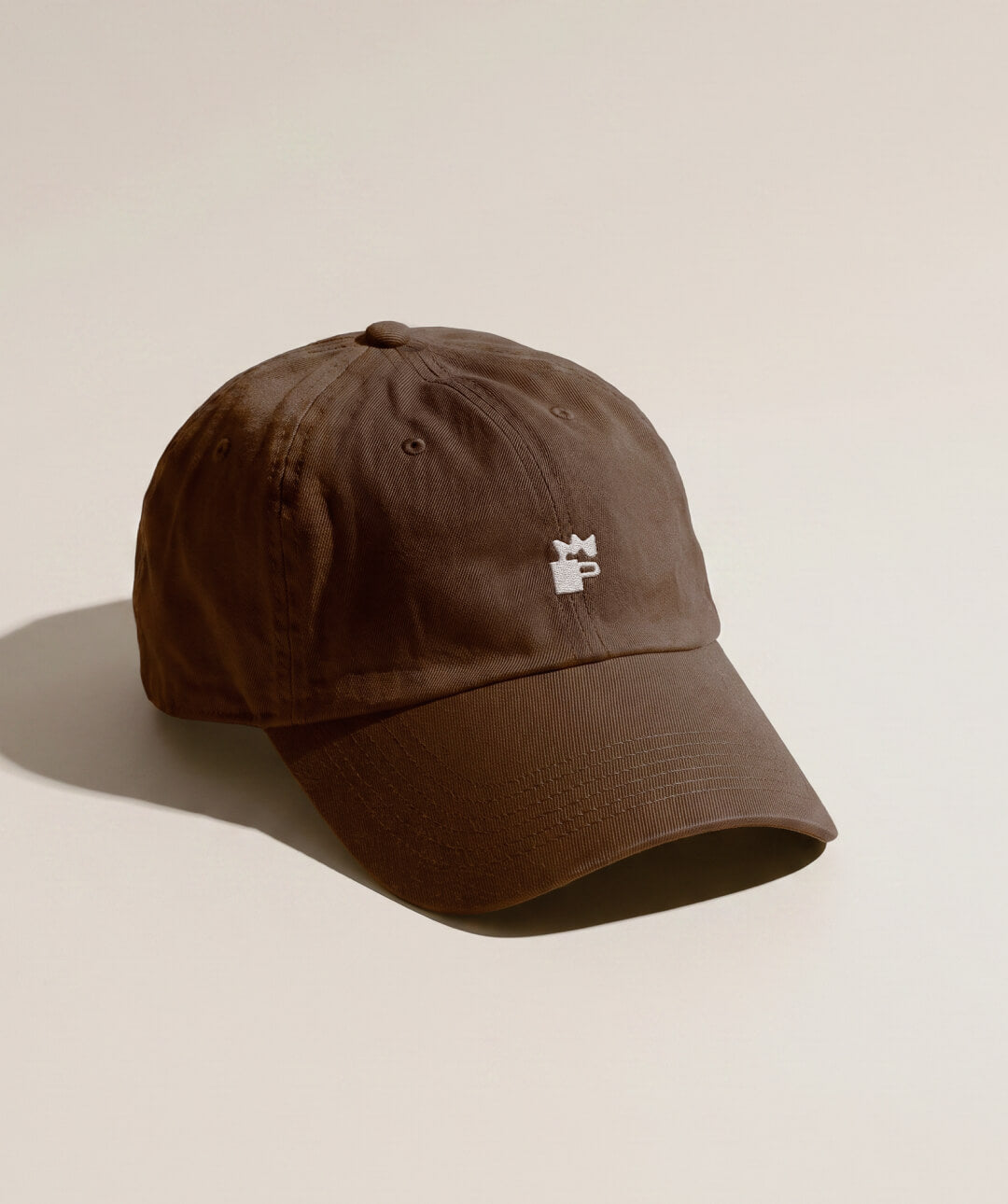 A brown cotton baseball cap with a small white NEIGHBOR logomark embroidered logo on the front, photographed on a warm beige background with a soft shadow.
