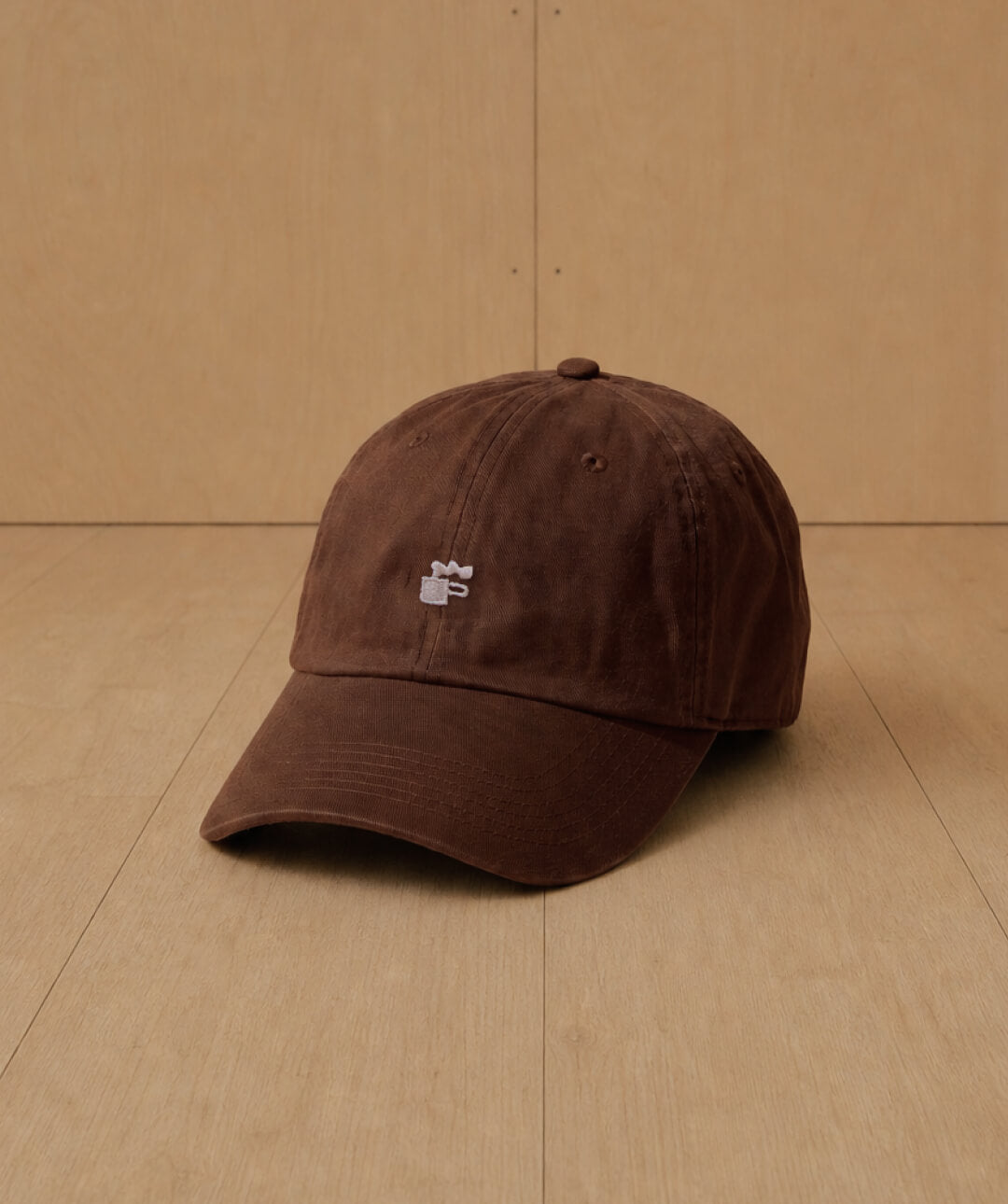 A dark brown cotton baseball cap with a small white NEIGHBOR Coffee logomark embroidered logo on the front, resting on a light wood floor against a matching wood panel background.