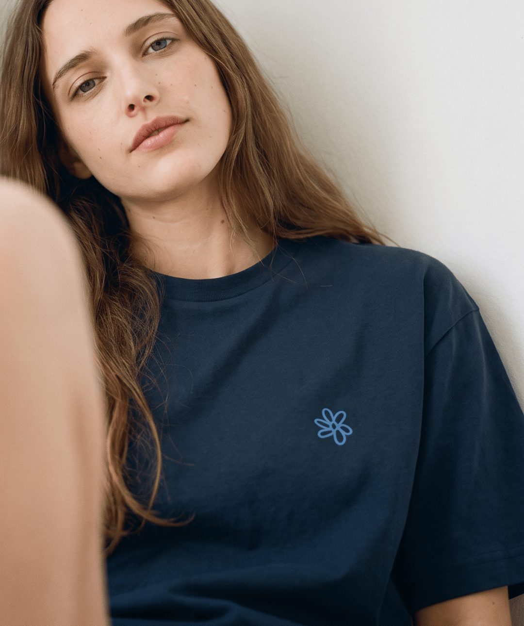 A close-up of a woman wearing a dark navy blue t-shirt with a small light blue embroidered flower on the left chest, leaning against a plain light wall.