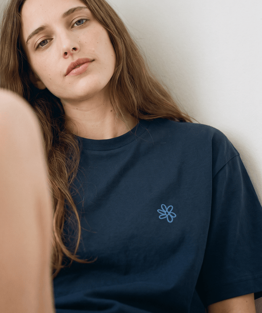 A close-up of a woman wearing a dark navy blue t-shirt with a small light blue embroidered flower on the left chest, leaning against a plain light wall.
