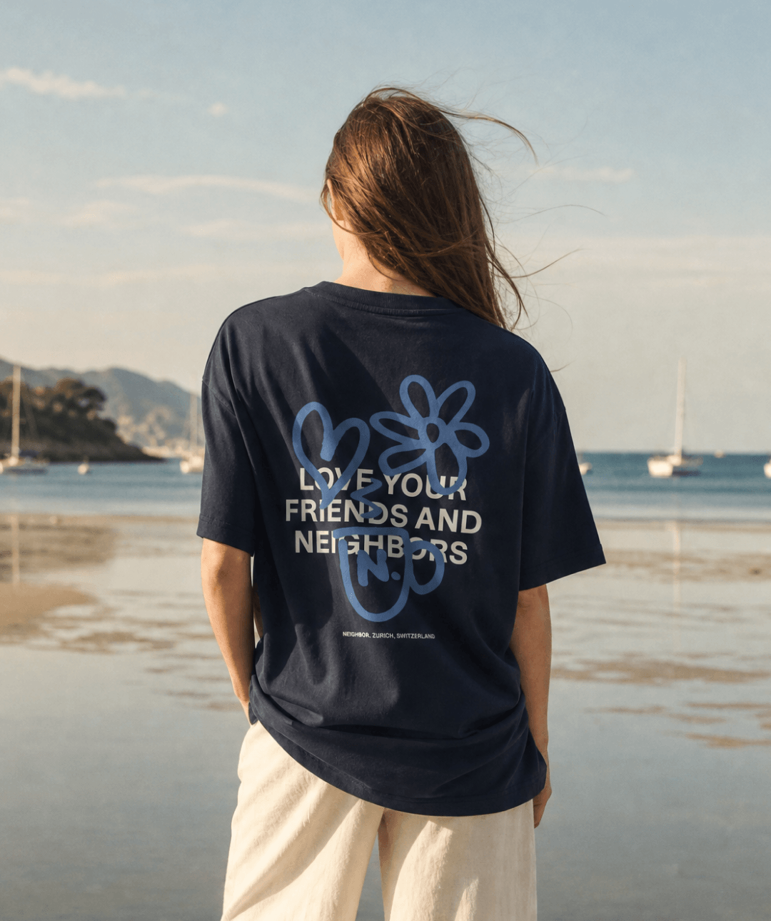A woman stands on a beach with her back to the camera, wearing a navy blue graphic t-shirt with a blue and white flower design and the text 'LOVE YOUR FRIENDS AND NEIGHBORS' on the back.