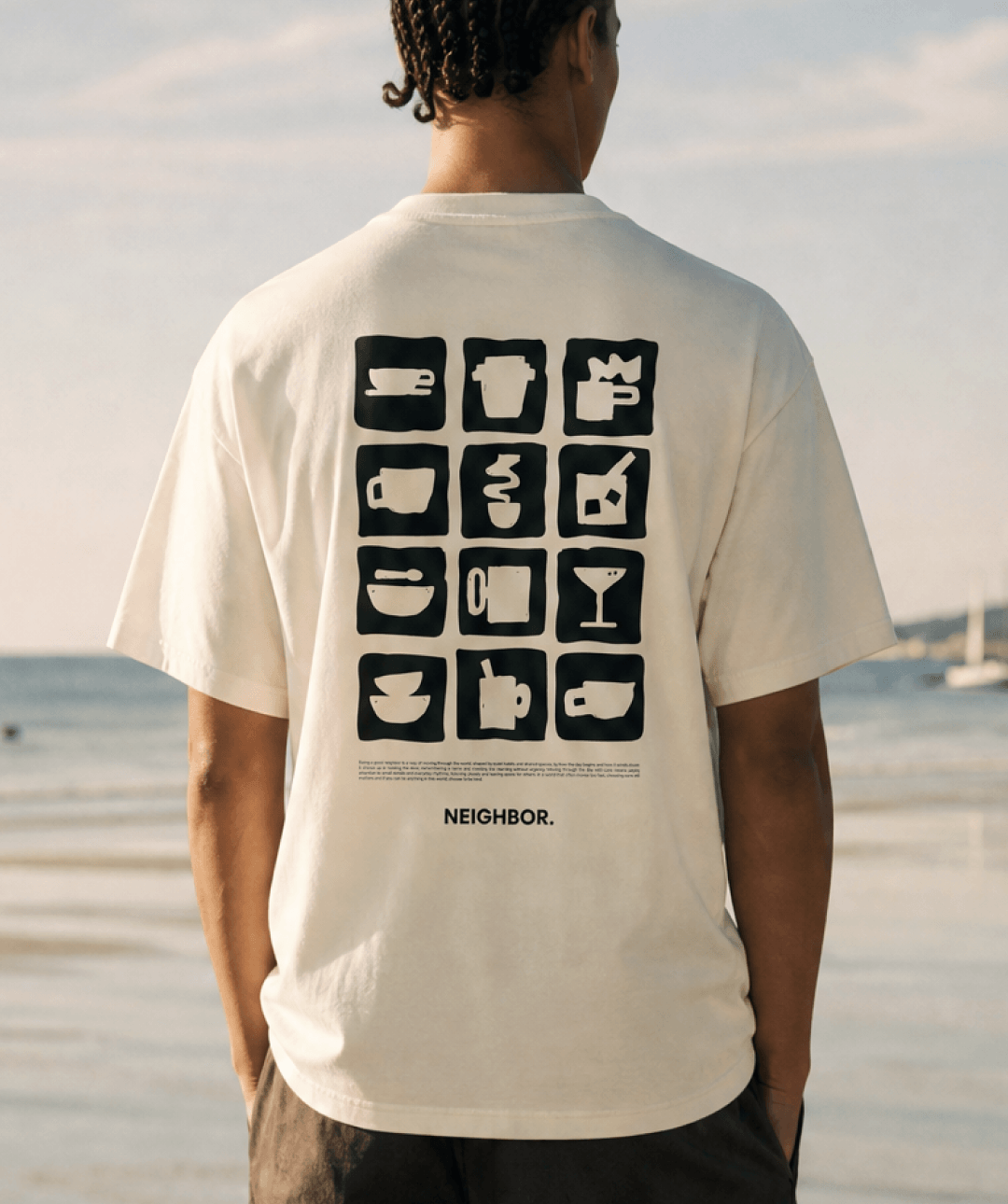 A man viewed from behind wearing a cream-colored t-shirt with a black graphic of 16 coffee cup icons and the word 'NEIGHBOR.' printed below, set against a beach background.