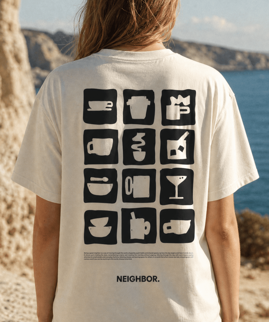 A person viewed from behind wearing a cream-colored t-shirt with a black graphic print of coffee-themed icons and the word 'NEIGHBOR.' on the back, set against a beach and ocean background.