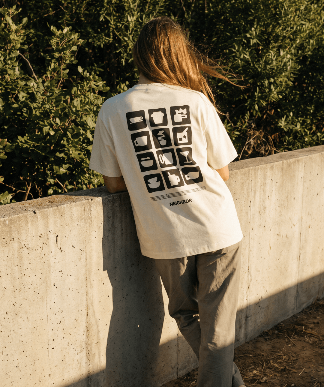 A woman viewed from behind wearing a cream-colored t-shirt with a black graphic of 16 coffee cup icons and the word 'NEIGHBOR.' printed below, set against a cement wall background.