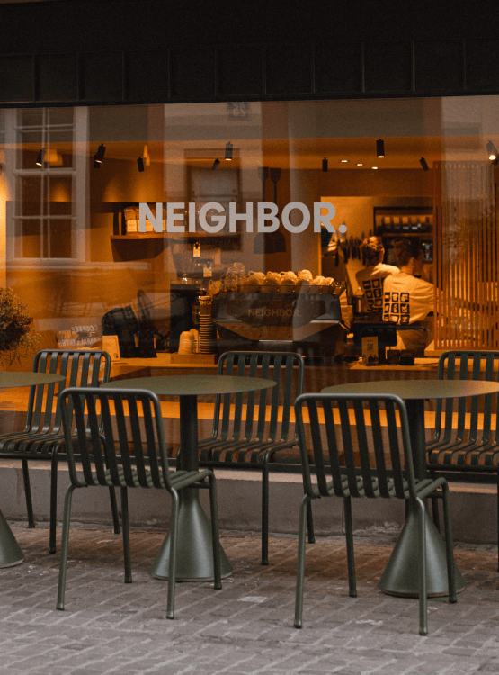 Outdoor cafe seating at NEIGHBOR with dark green metal tables and chairs set in front of a large window displaying a warm-toned interior. 