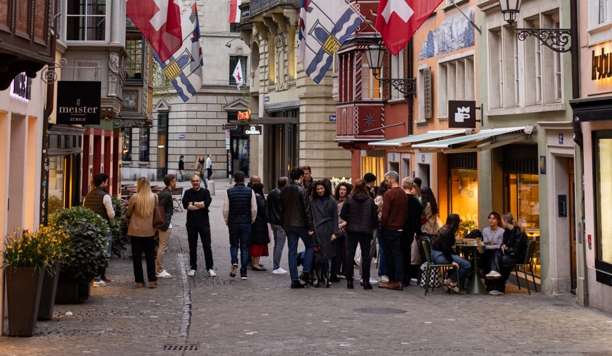 A narrow Zurich street lined with buildings and many Swiss flags hanging from balconies and storefronts. Pedestrians walk along the cobblestone street, and outdoor seating for NEIGHBOR cafe is visible on the right.