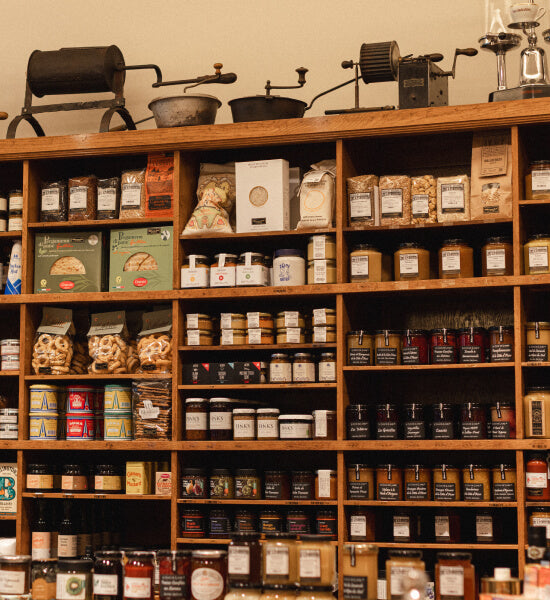 Floor-to-ceiling wooden shelving inside Schwarzenbach's Zurich shop, filled with jars of spices, tins, specialty food products, crackers, and coffee bags. Antique coffee grinders and roasting equipment line the top shelf.