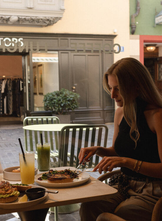 A woman sits at NEIGHBOR cafe in Zurich table cutting a plate of food, with drinks and other tables visible in the background.