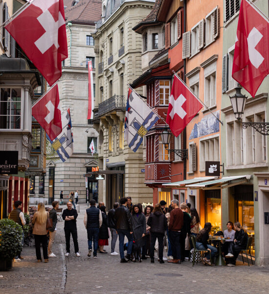 A narrow Zurich street lined with buildings and many Swiss flags hanging from balconies and storefronts. Pedestrians walk along the cobblestone street, and outdoor seating for NEIGHBOR cafe is visible on the right.